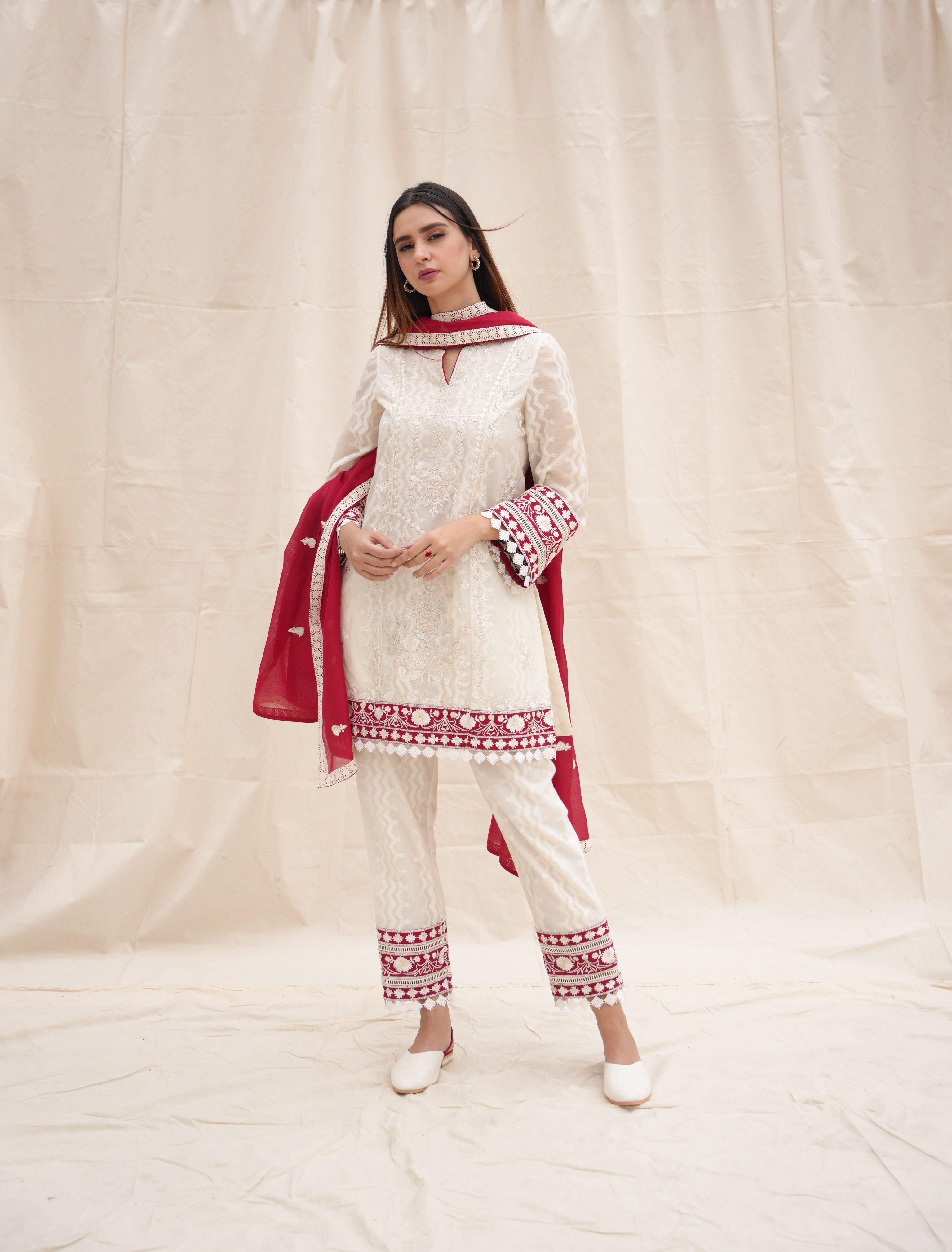 A woman poses in an elegant white traditional giza cotton outfit with red embroidery. She wears a matching red chanderi dupatta and white shoes, standing against a plain backdrop.