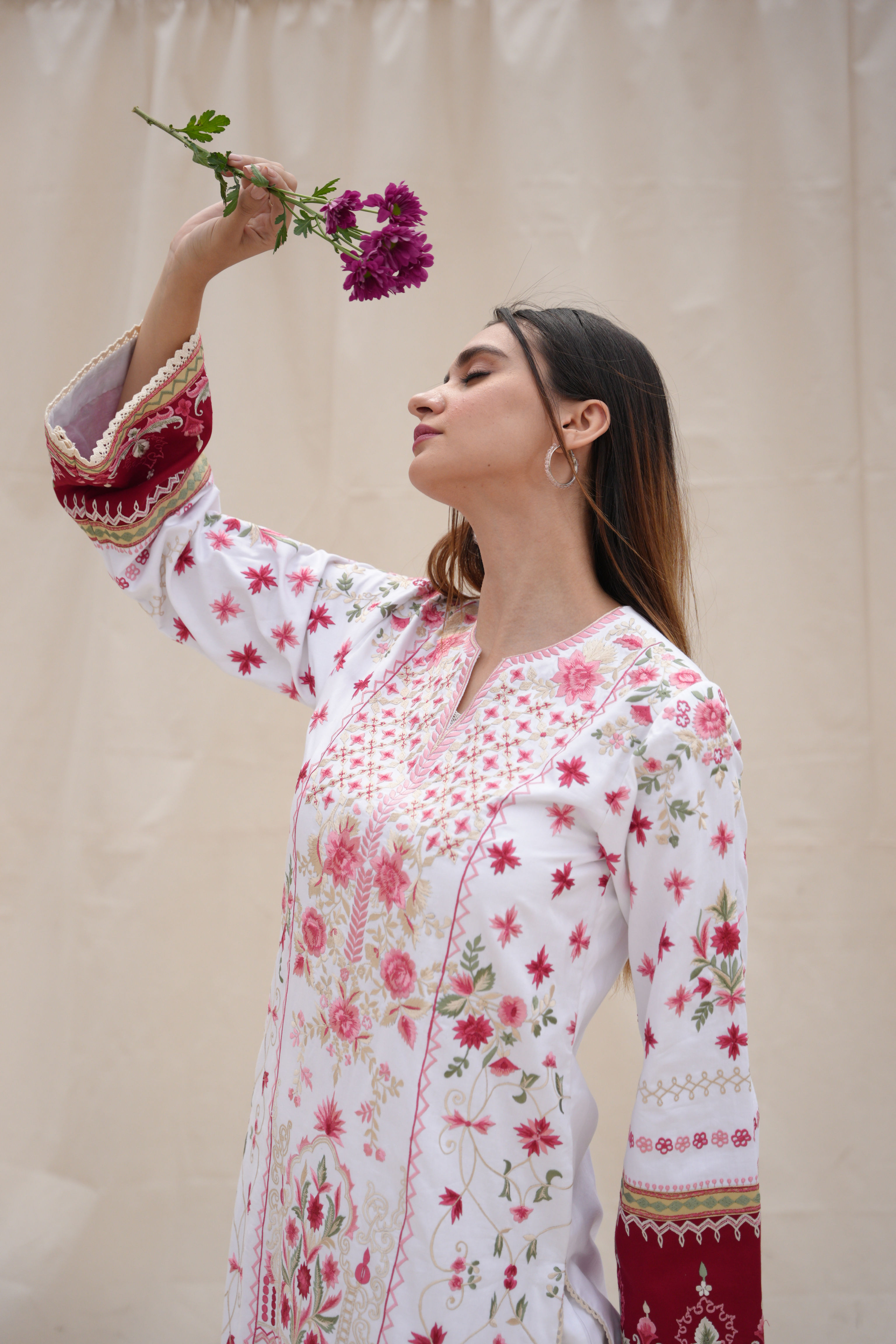 A woman in a floral dress with intricate pink and red embroidery design holds a purple flower up, eyes closed, exuding a serene and contemplative mood.