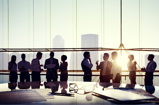 Silhouetted people conversing in an office with large windows. Sunlight filters in, illuminating papers and glasses on a table.