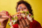 Woman with festival glitter make up and face paint and red nails holding a loaded taco, smiling against a bright yellow background.
