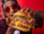 Person with festival glitter makeup holding a large burger with cheese and greens on a red background. The mood is playful and vibrant.