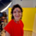 Woman in a red shirt with colorful festival glitter makeup, face paint and beaded necklace smiles in a studio. Yellow backdrop, camera, and light visible.