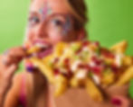 Woman with colourful festival glitter face paint joyfully eating loaded fries. Green background, vibrant colours, festive mood.
