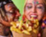 Two people with vibrant festival glitter face paint and accessories, sharing loaded fries.