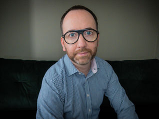 Portrait of an easy-to-talk-to online counsellor and psychotherapist wearing glasses and a checkered shirt, sitting on a dark couch in a calm, neutral setting.