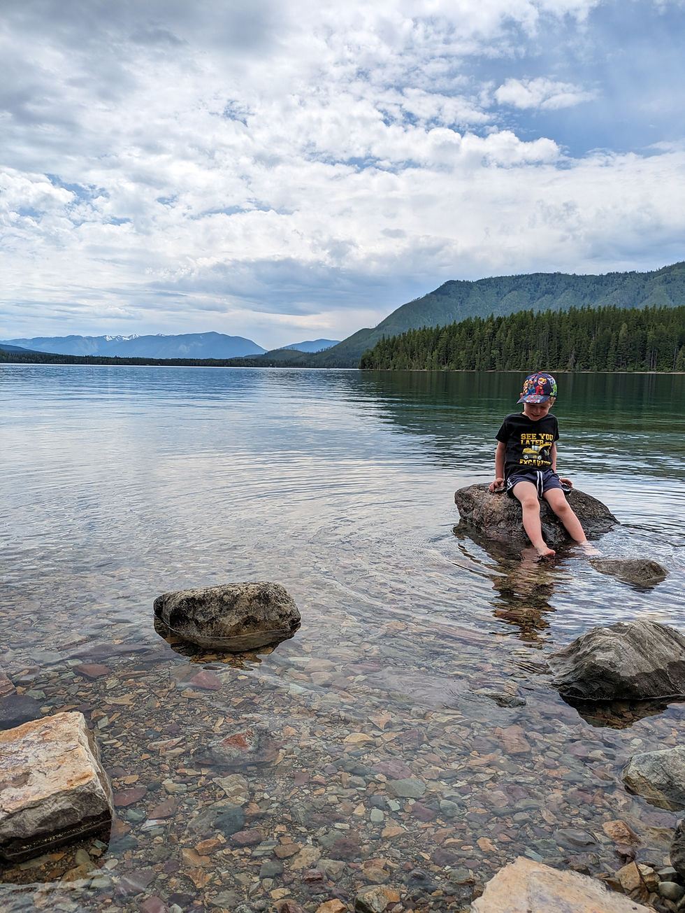 Child sitting on a rock in the shallow, crystal-clear waters of Lake McDonald, with forested mountains and a cloudy sky in the background.