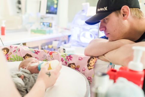 perth-birth-photographer-captures-prematurely-born-baby-boy-born-at-24-weeks-laying-in-his-crib-drinking-a-bottle-of-milk-with-a-dora-the-explorer-blanket-and-dad-looking-at-baby-the-perth-nicu-photography-project