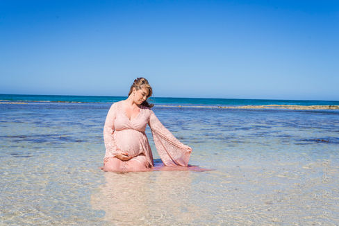 pregnant-mum-to-be-sitting-in-water-at-fishermans-hollow-beach-in-yanchep-holding-her-bump-in-a-pink-lace-dress-hired-from-bumps-that-borrow-beautiful-beginnings-perth-maternity-photographer