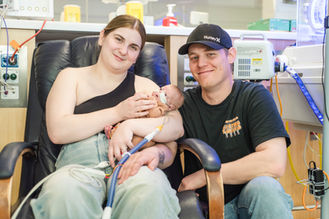 perth-birth-photographer-captures-new-mum-and-dad-holding-prematurely-born-son-in-king-edward-memorial-hospital-baby-boy-wearing-an-orange-jumpsuit-connected-to-high-flow-oxygen-perth-nicu-photography-project