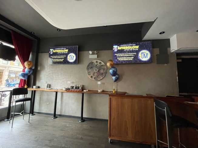 A bar along a wall with some small table-top balloon bouquets. Two landscape oriented screens are mounted on the wall above the bar with the happy hour logo and text that says "When it's time for you to "phone home", we are here to answer the call. We are grateful to be a part of your journey home".