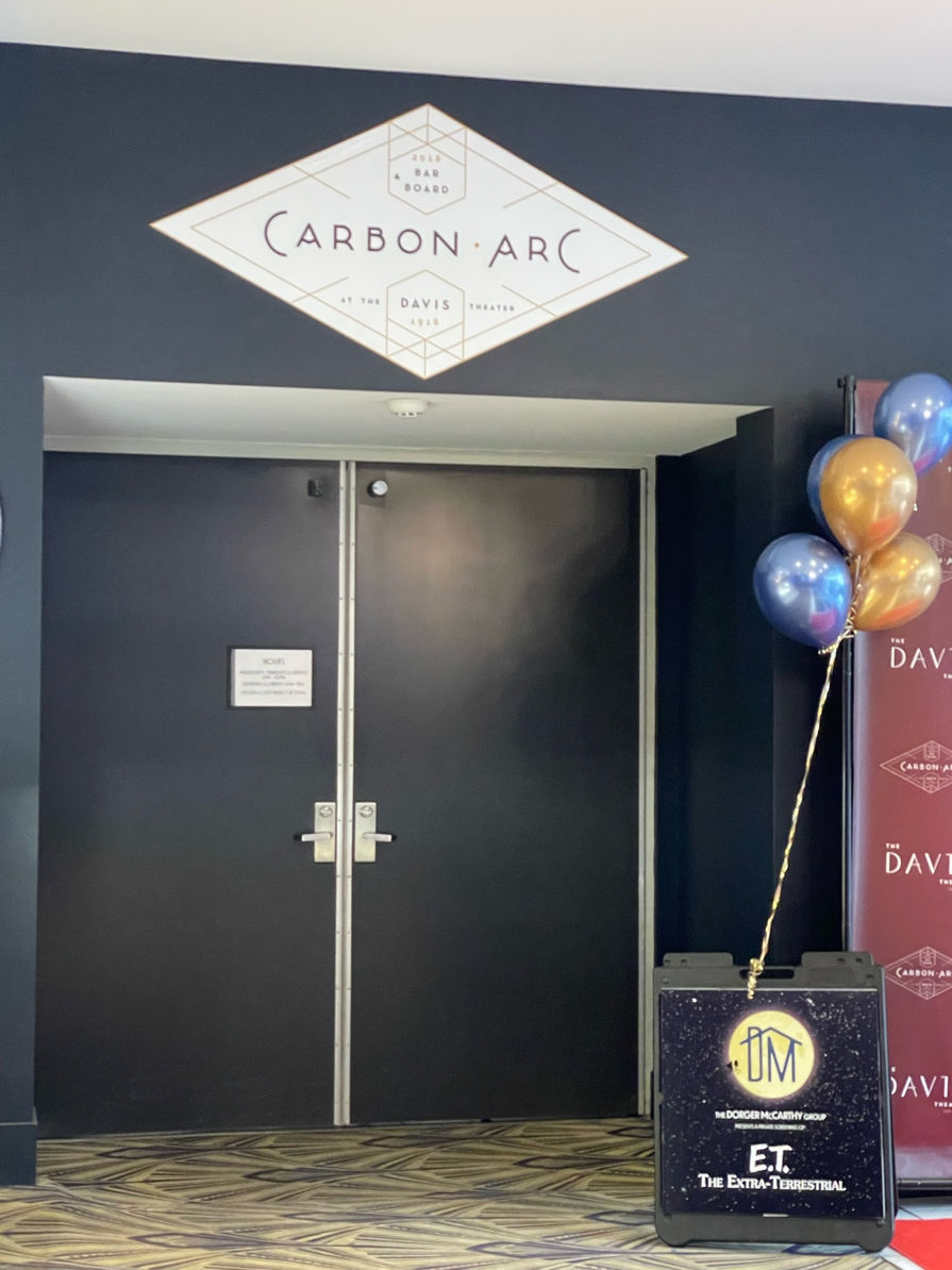 Black double doors with a sign above that reads "Carbon Arc". A sandwich board sign with the event logo stands to the right of the doors and has a bouquet of balloons tied to the top of the board.
