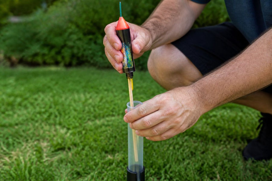 Close-up of a bottle rocket being inserted into a clear plastic launch tube in an outdoor backyard setting with grass visible, showing the stabilizing stick aligned vertically and the fuse at the top ready for ignition