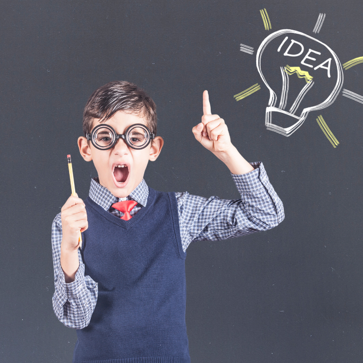 Boy in glasses excitedly points up, holding a pencil. He stands against a chalkboard with a drawn light bulb labeled "IDEA." Enthusiastic mood.