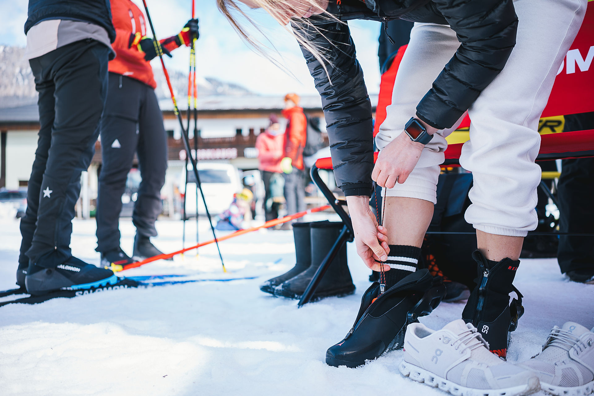Frau zieht Atomic Klassik Langlaufschuhe an im Hintergrund sind Salominski und Leki Langlaufstöcke zu sehen