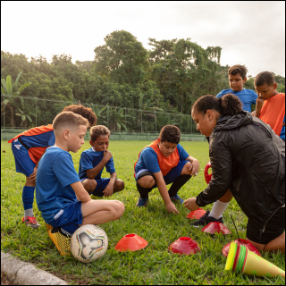 Children in blue soccer uniforms gather around a coach on a grassy field, focused on setting up orange cones. Trees line the background.