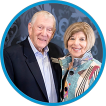 Older smiling man with gray hair in a dark suit standing with an attractive older woman with frosted blond hair and a patterned jacket.