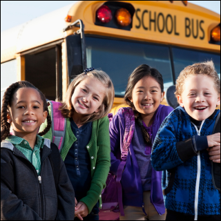 Four smiling children with backpacks stand in front of a yellow school bus. The sign on the bus reads "School Bus." Bright, cheerful mood.