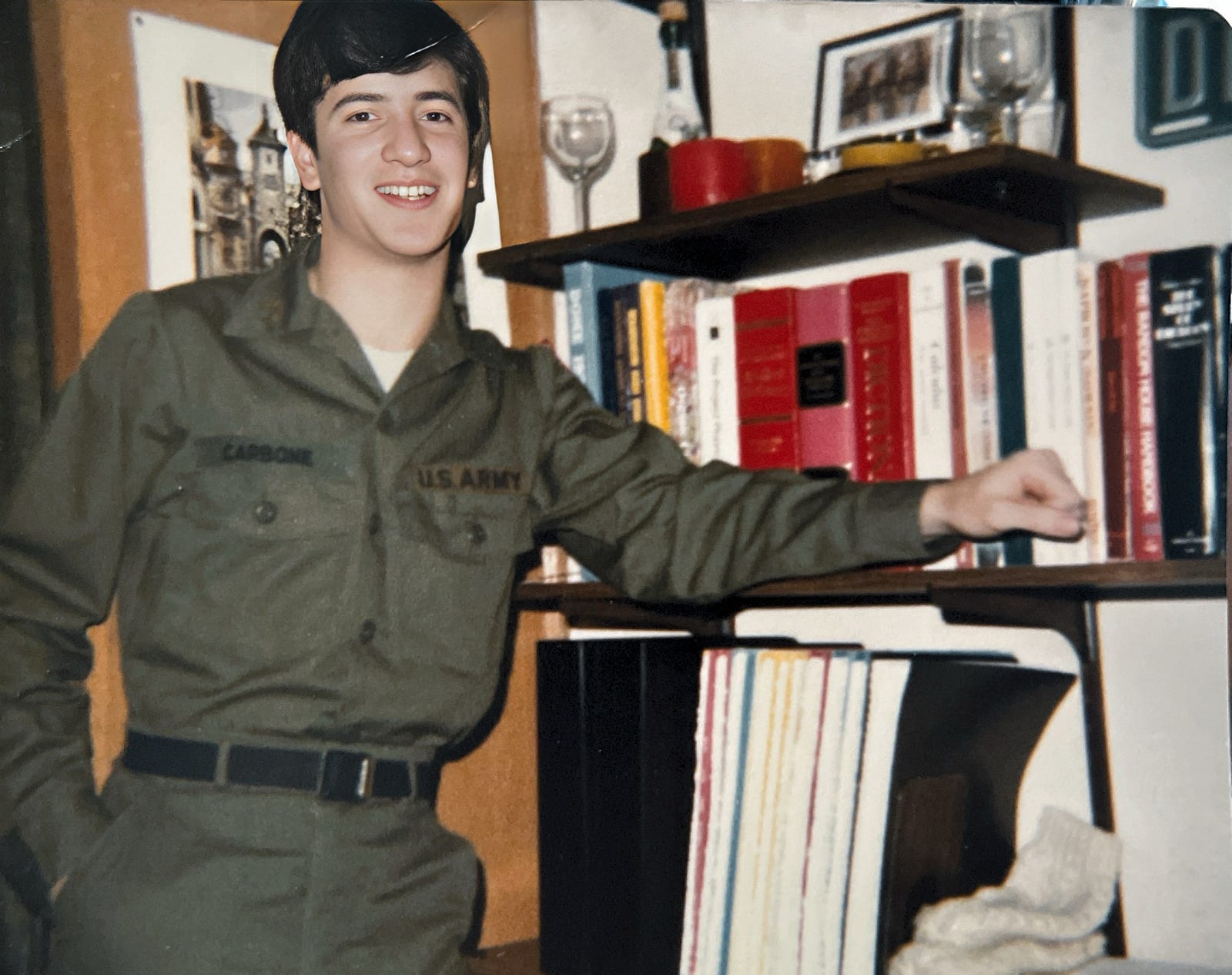In my Army ROTC fatigue uniform in my room at Fisher Hall, University of Notre Dame. Autobiography of Dr. Anthony J. Carbone. [Young Army ROTC cadet wearing olive drab fatigue uniform. In his college dormitory room. Floating shelves on wall filled with text books, notebooks, candles and souvenirs from Germany.]