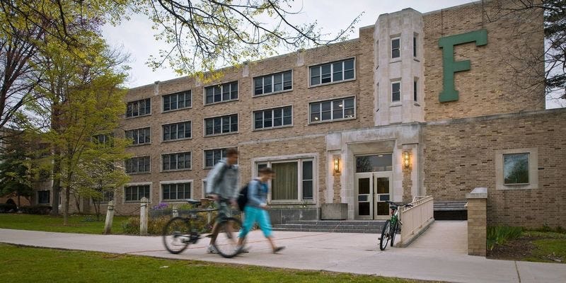 Photograph of Fisher Hall on the South Quad campus of the University of Notre Dame. With the famous big green “F” for Fisher on the front. Two students walking by, one holding onto his bicycle as they walk past the dorm. Autobiography of Dr. Anthony J. Carbone.