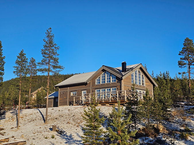 Brun stuga med snötäckt tak, träd, molnfri blå himmel. Fjällstugebyggarna.