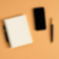 A flat lay image showing a black smartphone, a spiral-bound notebook with a pen tucked into the spine, and a black and gold fountain pen, all arranged on a light orange background.