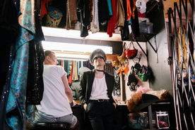 Two people are getting ready for a show backstage. There are costumes hanging around them. The person on the left is sitting facing a mirror with their head turned toward the other person. The person on the right leans against the counter behind them and faces the camera. 