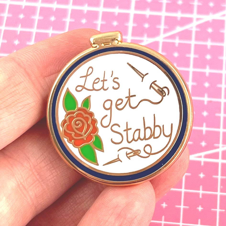 Thumbnail: A magnetic enamel needle minder with a metallic gold-tone edge, featuring a red rose and the phrase 'Let's Get Stabby' on a w