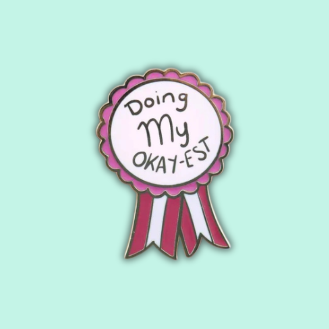 A pink and white enamel lapel pin with the text 'Doing My Okay-est' on it, designed in a ribbon style.
