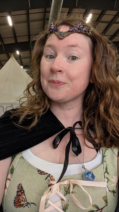 Alicia smiled in a selfie. She wears a crown in her curled red hair. Her top is a green lace up one, and she has a black cape on her shoulders as she's at a Ren Faire