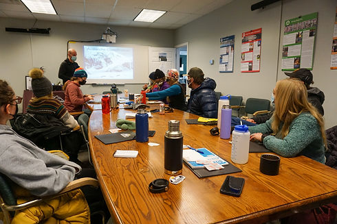 Students attending an indoor avalanche safety lecture with maps and materials on the table.