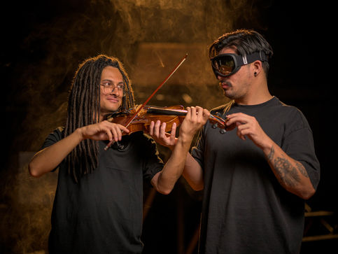 Un hombre de pelo largo toca un violín mientras otra persona que usa un visor toca el violín para sentir la vibración de la música. Hay humo en el ambiente y una luz cálida.
