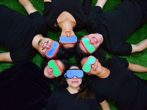 Dos hombres y cuatro mujeres están acostados en el piso formando un círculo con sus cabezas. Todos usan visores que tapan sus ojos y camisas negras.