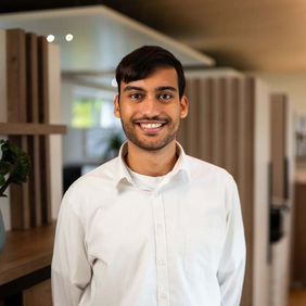 Smiling man in white shirt, standing in an office, looking at camera.