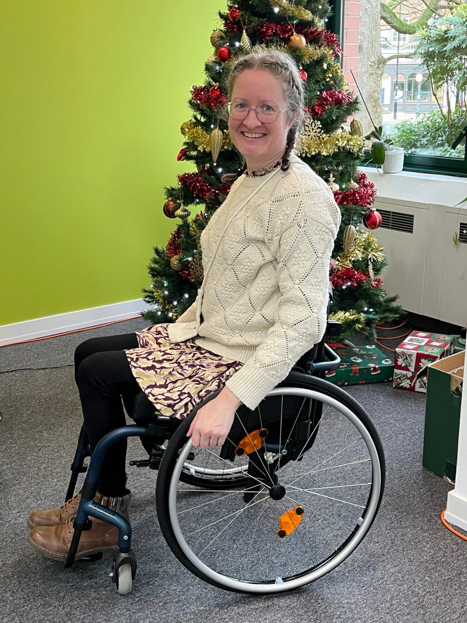 white woman with glasses and brown hair, sat in a wheelchair that has midnight blue frame, and looks rather smart.