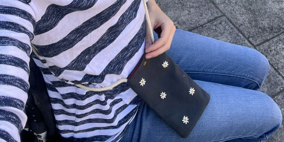 seated person wearing a striped top and blue jeans, with a navy blue phone bag embroidered with daisies on their lap