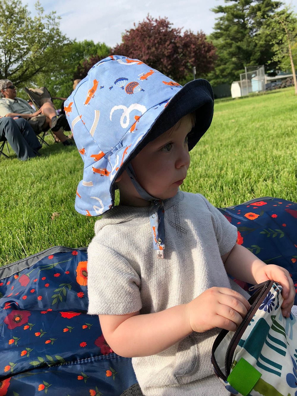 Caucasian toddler sitting on a blanket outdoors, looks off in the distance. He is wearing a blue bucket hat with airplanes printed on it, and holding a reusable bag in his hand. He is sitting on a blue floral blanket with red and orange flowers printed on it. In the background is grass, trees, and people sitting in lawn chairs.