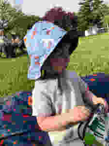 Caucasian toddler sitting on a blanket outdoors, looks off in the distance. He is wearing a blue bucket hat with airplanes printed on it, and holding a reusable bag in his hand. He is sitting on a blue floral blanket with red and orange flowers printed on it. In the background is grass, trees, and people sitting in lawn chairs.