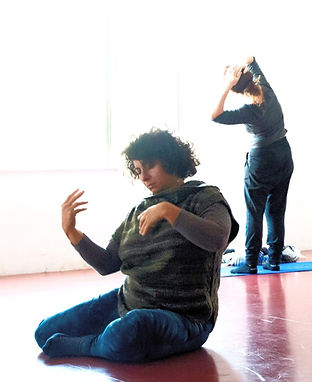 Two women practicing Authentic Movement, one standing and inclining by a window and the other sitting on the floor, embodying self-exploration and connection in a group session.