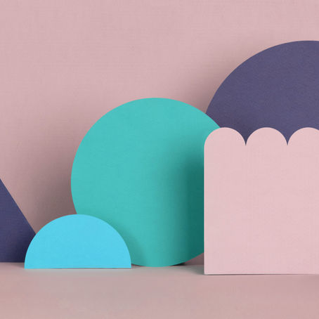 Colorful geometric paper shapes on a pastel pink background. Shapes include blue circles, a triangle, and a scalloped pink rectangle.