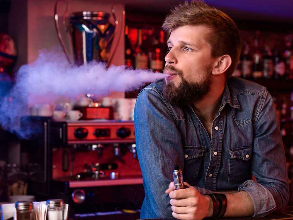 A bearded man in a denim shirt exhales a cloud of vapor from an e-cigarette while leaning against a bar or café counter.