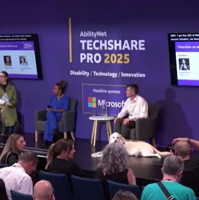 Panel at AbilityNet TechShare Pro 2025 on disability tech, with sign-language interpreter, seated speakers Samanta Bullock, Founder of Bullock Inclusion, Maxine Williams, VP Head of Accessibility & Engagement at Meta, Robin Christopherson, Head of Digital Inclusion at AbilityNet, audience, and service dog.