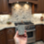 Hand holding a granite sample in a kitchen, matching the brown cabinets and beige speckled backsplash. Stove and soft lighting in background.