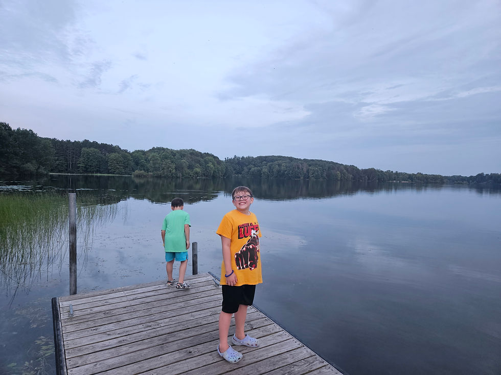 Two kids stand on a dock by a calm lake. One wears a yellow shirt and smiles, while the other looks at the water. Trees line the horizon.