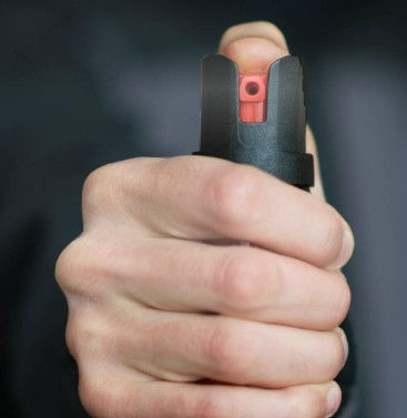 Hand gripping a black and red pepper spray canister, pointing forward. Blurred dark background, conveying a sense of alertness or caution.