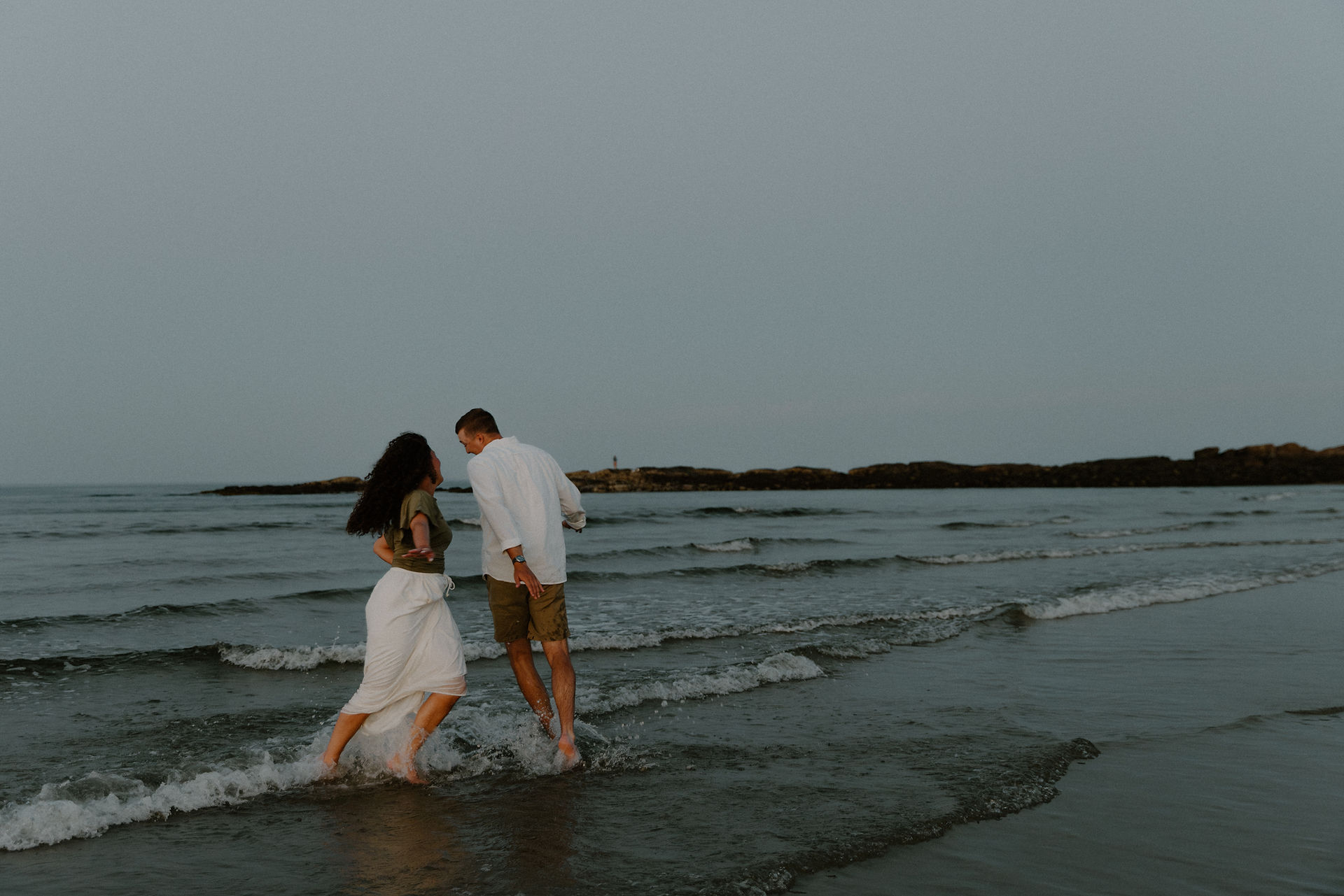 Maine couple chase each other in the water during sunset at beach during coastal engagement session