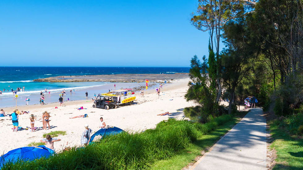 People relaxing on a sunny beach, some swimming. A yellow vehicle is parked near the shore. Green grass and trees line a nearby path.