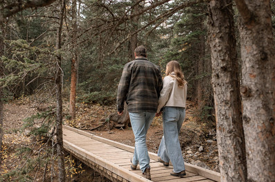 Couple walking hand in hand along a wooden forest path during Sheep River engagement photos, surrounded by tall trees and a peaceful natural setting.