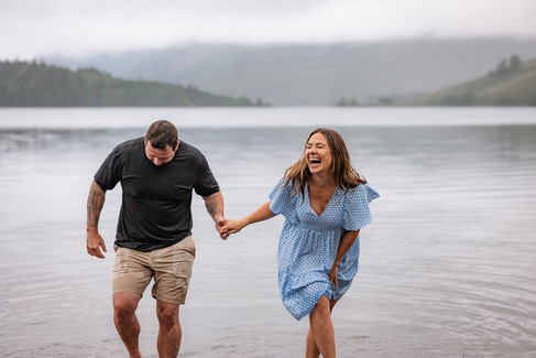 Couple laughing and holding hands while running through the water during fun engagement photos captured by a Waterton Park wedding photographer.