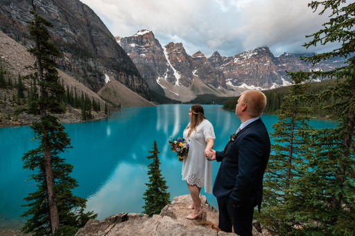 Bride and groom standing on big rocks holding hands looking out onto Moraine Lake.