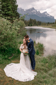 Bride and groom embracing beside a mountain lake during their Canmore Nordic Centre wedding, with breathtaking Rocky Mountain peaks and lush evergreen trees in the background.